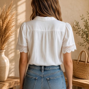 May include: A white short-sleeved blouse featuring a classic collar and delicate lace trim detailing on the sleeve cuffs, paired with blue denim jeans. The blouse has a button-down front and a structured back yoke with a subtle vertical pleat.