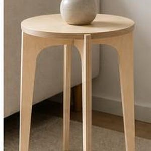 May include: A round light-colored wooden side table with three legs featuring arched cutouts. A small, smooth, light gray ceramic vase sits on the tabletop, containing a green leafy plant with multiple stems and small oval-shaped leaves.