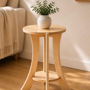 Puede incluir: Una mesa auxiliar de madera clara con una superficie circular y un estante inferior circular más pequeño. La mesa cuenta con tres patas curvas que se extienden desde la parte superior hasta la base. Un jarrón de cerámica blanca con una planta verde descansa sobre la mesa.