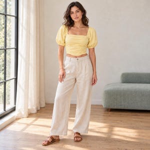 May include: A pale yellow cropped blouse with puff sleeves and a square neckline paired with high-waisted, wide-leg beige linen trousers. The outfit is completed with simple brown leather slide sandals, creating a coordinated ensemble.