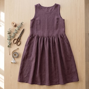 Puede incluir: Un vestido sin mangas hasta la rodilla en color ciruela oscuro sólido con cuello redondo y cintura fruncida. El vestido cuenta con bolsillos laterales y un corpiño sencillo. Junto al vestido hay tijeras de metal, un carrete y una cinta métrica.
