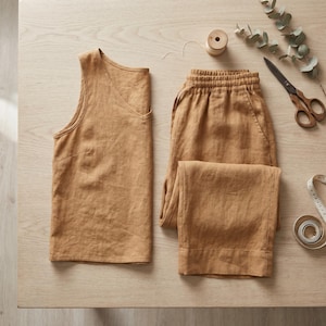 May include: A sleeveless top and matching elastic-waist trousers in a solid tan color. The set is displayed alongside a wooden spool, a pair of metal scissors, and a white measuring tape, showcasing the fabric texture and garment construction.