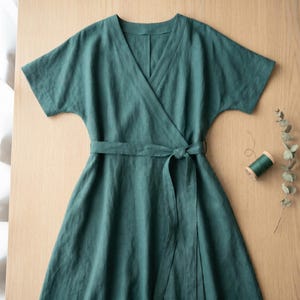 May include: A dark green wrap dress featuring short sleeves, a V-neckline, and a matching fabric tie belt at the waist. The garment is constructed with a visible weave texture and is displayed flat, accompanied by a small spool of green thread.