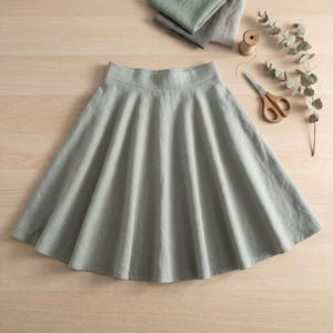 May include: A sage green flared skirt with a smooth waistband and a full, pleated silhouette. The skirt is displayed alongside a wooden spool of thread, a pair of metal scissors, and a sprig of dried eucalyptus leaves on a light wood surface.