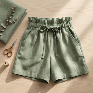 May include: Sage green shorts featuring a high-waisted paperbag style with an elasticated waistband, a matching fabric drawstring tie, and two side pockets. The shorts are displayed alongside a pair of metal scissors and folded fabric.