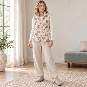May include: A cream-colored zip-up vest featuring a repeating blue floral pattern, worn over a long-sleeved white shirt and light beige trousers. The outfit is completed with a pair of light-colored slip-on shoes.