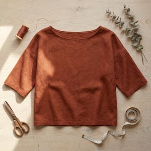 May include: A rust-coloured short-sleeve top with a wide boat neckline and a straight hem. The garment is displayed alongside a spool of thread, a pair of metal scissors, a measuring tape, and a sprig of eucalyptus leaves on a light wood surface.