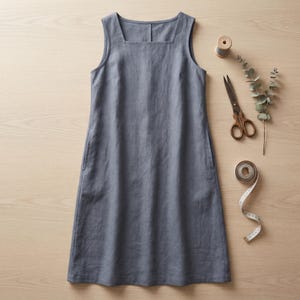 May include: A sleeveless slate blue dress with a square neckline and a straight silhouette. Beside the dress are a pair of metal scissors, a small wooden spool, a sprig of eucalyptus, and a white measuring tape with visible inch markings.