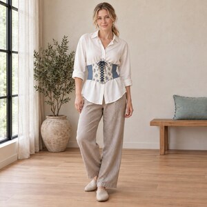 Puede incluir: Una camisa blanca de manga larga con botones, combinada con un corsé azul y blanco con estampado floral y cordones frontales, junto con pantalones beige de corte holgado y zapatos sin cordones de color claro.
