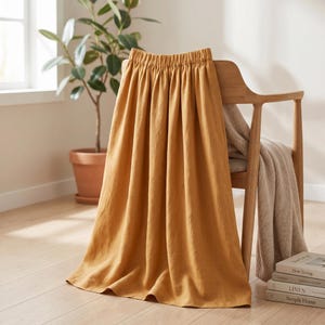 May include: A mustard yellow midi skirt with an elasticized waistband and a gathered, flowing silhouette. The skirt is draped over a wooden chair, showcasing its soft, textured fabric and deep, warm color tone in a bright, indoor setting.