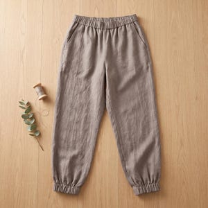 May include: Taupe-colored trousers featuring an elasticized waistband and elastic cuffs at the ankles. The pants have two side pockets and a relaxed fit, presented alongside a small wooden spool of matching thread and a sprig of eucalyptus.