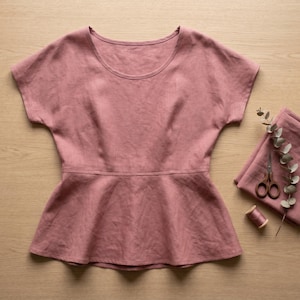 May include: A dusty rose-colored short-sleeve top with a round neckline and a horizontal seam at the waist that creates a flared peplum hem. The garment features a simple design with short sleeves and a fitted bodice that transitions into a wider skirt.