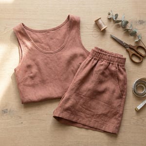May include: A dusty rose sleeveless tank top and matching elastic-waist shorts set. The garments feature a simple design with a scoop neckline on the top and side pockets on the shorts, presented alongside sewing tools including scissors and thread.
