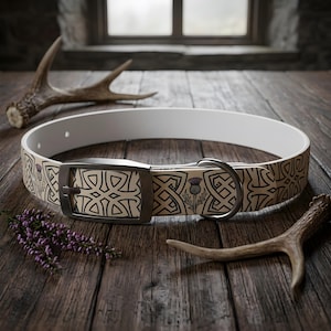 May include: A dog collar featuring a cream-colored strap with a black Celtic knot pattern and purple thistle illustrations. The collar includes a dark metal buckle and a D-ring, with a white interior lining, presented on a dark wooden surface.
