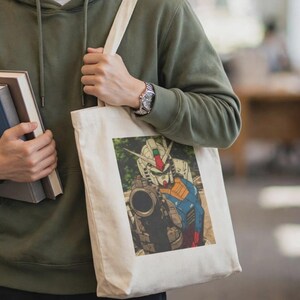 以下が含まれることがあります： ドローストリング付きのオリーブグリーンのパーカーと、大きな武器を持ったロボットのキャラクターのグラフィックプリントが施された白いキャンバストートバッグ。トートバッグは肩にかけられ、手にはハードカバーの本が2冊持たれています。
