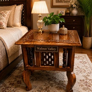 Puede incluir: Una mesa auxiliar cuadrada de madera con acabado marrón oscuro, con bordes tallados intrincados en la superficie superior, cuatro patas curvas y un panel central de celosía. Una placa rectangular muestra el texto Walnut Valley.
