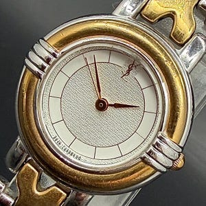 May include: A two-tone wristwatch featuring a round silver and gold-colored bezel with a textured white dial. The watch face displays thin black hour markers, three slender hands, and the YSL logo in the upper right quadrant of the dial.