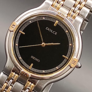 May include: A Seiko Dolce wristwatch featuring a round black dial with gold-tone hands and hour markers. The bezel displays Roman numerals at the twelve, three, six, and nine o'clock positions. It has a two-tone silver and gold metal link bracelet.