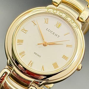 May include: A gold-tone Seiko Lucent wristwatch featuring a round white dial with gold Roman numeral hour markers. The watch has gold hands and a matching gold-tone metal link bracelet. The word LUCENT is engraved on the top bezel and printed on the dial.
