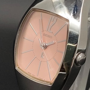 May include: A Seiko Lukia wristwatch featuring a tonneau-shaped silver-tone case and a soft pink sunray dial. The dial displays silver-tone hour markers, a prominent number 12, silver hands, and the Seiko and Lk logos in the center.
