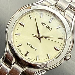 Könnte beinhalten: Eine silberfarbene Seiko Exceline Armbanduhr mit einem runden, champagnerfarbenen Zifferblatt, silbernen Stabindizes, drei zentralen Zeigern und einem polierten Metallgliederarmband. Das Zifferblatt zeigt das Seiko-Logo und den Schriftzug Exceline.