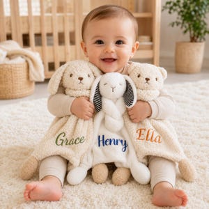 May include: Three plush security blankets featuring stuffed animal heads: a cream-coloured bunny with green embroidery reading Grace, a white bunny with black and white striped ears and blue embroidery reading Henry, and a cream bear with orange embroidery reading Ella.