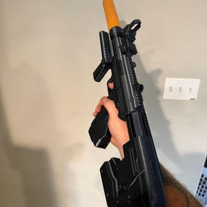 May include: A black toy submachine gun featuring a prominent orange barrel tip, a tactical rail system, a top-mounted sight, and a bulky rear stock. The toy weapon has a textured grip and a detachable magazine, constructed with a matte black finish.