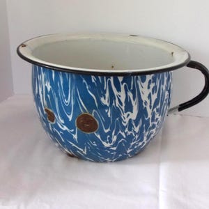 May include: A vintage enamelware chamber pot with a blue and white swirl pattern. The pot has a white interior, a black handle, and a black rim. Two brown spots are visible on the front.