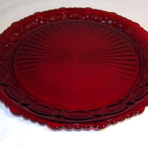 May include: A round, ruby red glass serving plate. The plate has a scalloped edge with a decorative pattern and a central design of radiating lines. The plate is likely vintage and suitable for serving food.