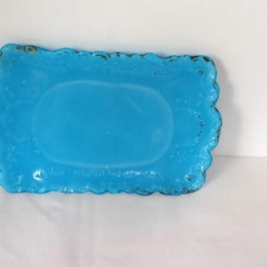 May include: A rectangular, turquoise-colored glass dish with a scalloped edge. The dish has a slightly raised oval center and a distressed, dark rim. The overall design is vintage-inspired.