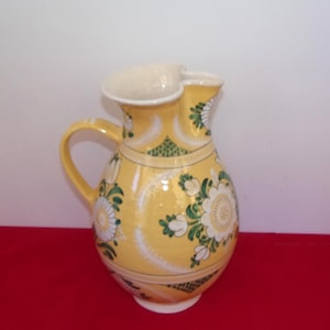 May include: A yellow ceramic pitcher featuring a white floral pattern with green leaves and geometric accents. The vessel has a curved handle, a wide spout, and a rounded body, decorated with white circular motifs and green cross-hatched details.