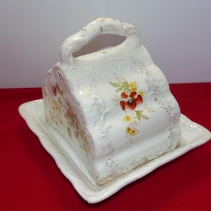 May include: A white ceramic butter dish with a handle, featuring floral designs in red, yellow, and green. The dish sits on a matching square plate with a scalloped edge. The overall design is ornate and vintage.