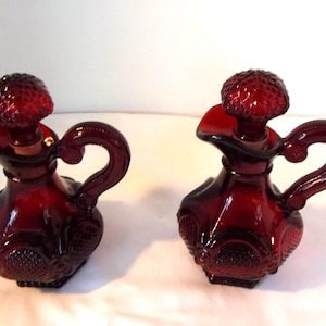 May include: Two ruby red glass cruet bottles with a textured pattern and a handle. Each bottle has a matching stopper. The bottles are likely for oil and vinegar and are displayed on a white surface.