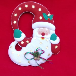 May include: A festive Christmas decoration in the shape of Santa Claus. The figure is primarily red and white, with gold star accents and green holly details. The Santa Claus figure is set against a solid red background.