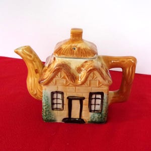 Peut inclure: Théière en céramique en forme de cottage. Elle présente un toit de chaume beige, une cheminée et une façade de maison avec une porte et des fenêtres. La poignée et le bec sont beiges. La théière est sur une surface rouge.