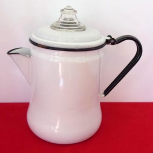 May include: White enamel coffee pot with a black handle and rim. The pot has a clear glass knob on the lid and a spout for pouring. The pot is sitting on a red surface.
