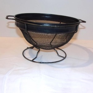 May include: A black metal strainer with a mesh basket and a circular base. The strainer has two handles and is designed for straining liquids or rinsing food. The kitchen tool is likely used for cooking and food preparation.
