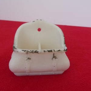 May include: Off-white, vintage wall pocket with a rounded top and a small hole for hanging. The pocket has a divided interior and a decorative, distressed metal trim. The base is rounded with vertical lines.