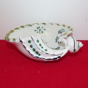 May include: A white ceramic bowl shaped like a seashell with a scalloped edge. The interior features green painted starburst and floral patterns. The exterior has a layered, fin-like structure with green dots and a metallic silver trim along the edges.
