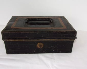 May include: A vintage black metal cash box with a rectangular shape. The box features a handle on the top and a lock on the front. The top has a brown border. The box is likely used for storing money or valuables.