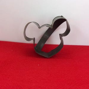 May include: A dark metal cookie cutter in the shape of a stylized bow or a Mickey Mouse head. The cutter is placed on a red surface with a white background.