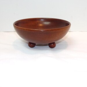 May include: A round, medium-brown wooden bowl with a smooth, polished finish. The bowl features a wide, open top and is supported by three small, spherical feet attached to the base, elevating the vessel slightly above the surface.