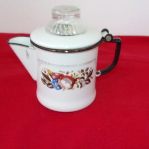 May include: White enamel coffee pot with a glass lid and black handle. The pot features a decorative floral and fruit design in red, blue, and brown. The spout is triangular, and the pot sits on a red surface.