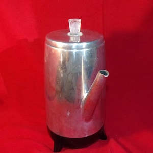 May include: A vintage, stainless steel coffee percolator with a clear glass knob on the lid. The percolator has a spout and sits on a black base. The background is a solid red.