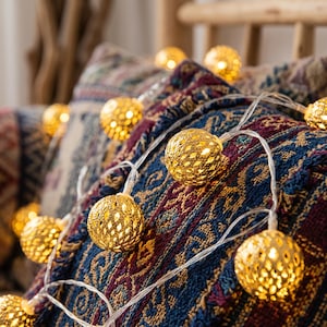 May include: A string of decorative lights featuring spherical gold-toned metal covers with a repeating diamond-shaped cutout pattern. Each sphere emits a warm glow from within, connected by a thin, clear electrical wire draped across a textured fabric.