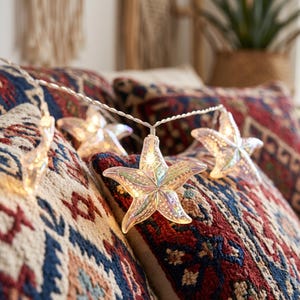 May include: A string of decorative lights featuring translucent, iridescent starfish-shaped covers. Each starfish has a textured surface that glows from an internal light source, connected by a thin, flexible white wire that drapes across pillows.