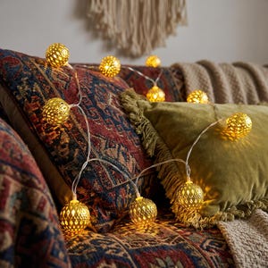 May include: A string of decorative lights featuring spherical gold-toned metal shades with intricate cut-out patterns. Each globe emits a warm, glowing light, connected by a thin, clear wire draped across textured fabric cushions and pillows.