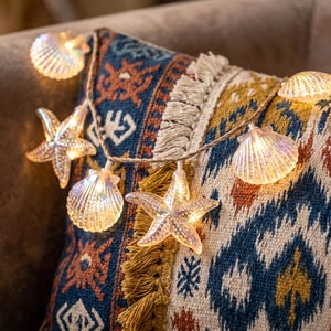 May include: A string of decorative LED lights featuring translucent, iridescent seashell and starfish shapes. The lights emit a warm glow, connected by a thin, twisted brown cord, creating a coastal-themed illumination piece for interior decor.