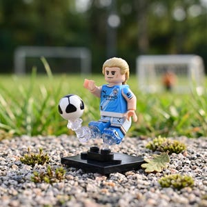 May include: A LEGO minifigure of a soccer player with blonde hair wearing a light blue Manchester City jersey with the Etihad Airways logo. The figure is posed mid-kick with a black and white soccer ball attached to a clear plastic stand.