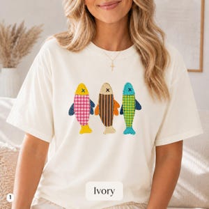 May include: An ivory short-sleeve t-shirt featuring a graphic of three stylised fish. The left fish has a pink and white gingham pattern, the middle fish has orange and navy vertical stripes, and the right fish has a green and white gingham pattern.